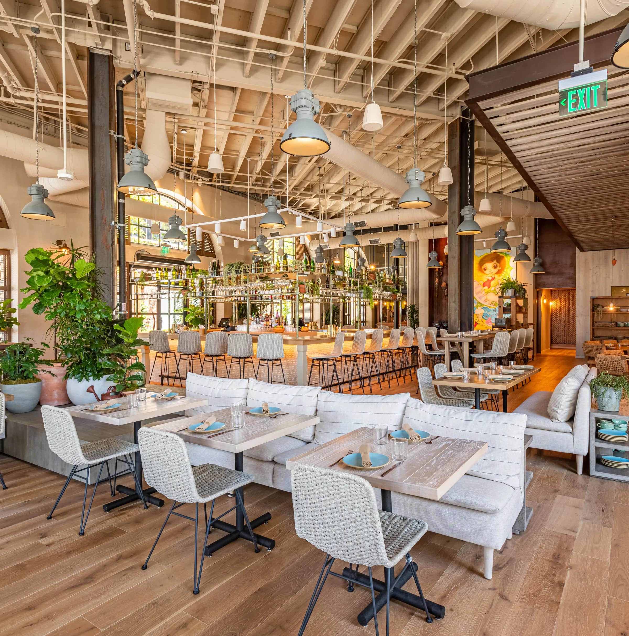 An airy restaurant dining room with a long bar, potted plants and set tables