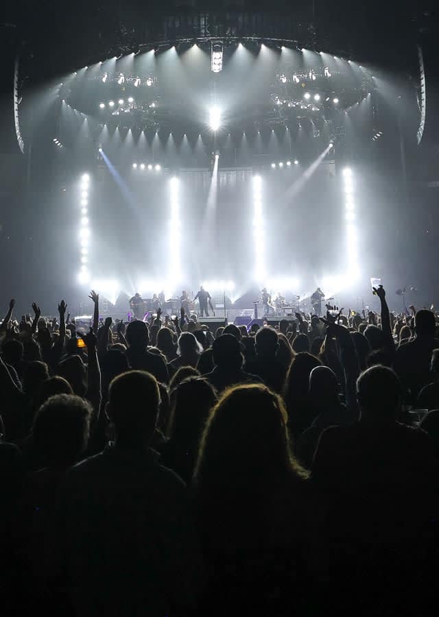 A packed arena crowd silhouetted against a brightly lit concert stage